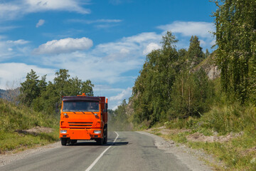 A Russian-made truck with an orange cab on the road outside the city on a summer day against the background of green trees and blue sky. Intercity delivery of construction cargo.