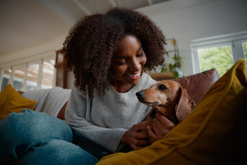 Smiling african woman playing with pet wiener dog at home sitting on couch