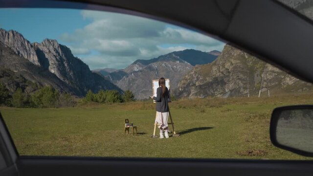 Beautiful girl paints a picture in the mountains. A professional artist creates a landscape on a molbert. Hobbies, hobbies and work in the fresh air.