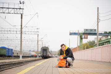 Naklejka na ściany i meble Young man squats on platform, waiting for train. Male passenger with backpacks sitting on railroad platform in waiting for train ride. Concept of tourism, travel and recreation.