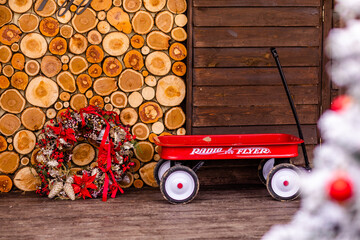 Christmas texture background of dry wooden log cabins and decorative wreath with cones, ribbons and berries and a red traditional cart