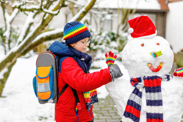 Little school kid boy in colorful clothes, with glasses and backpack having fun with snowman after...
