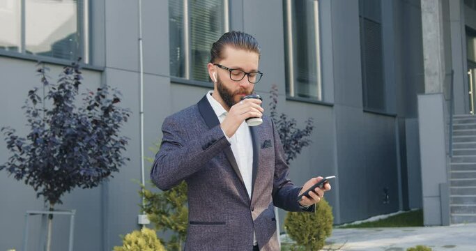 Side view of handsome confident concentrated bearded office manager which listening music in airpods from phone playlist and enjoying coffee during time break outdoors,4k