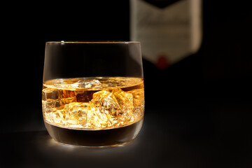 Whiskey with ice in glass in front of bottle on black background.
