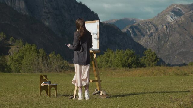Beautiful girl paints a picture in the mountains. A professional artist creates a landscape on a molbert. Hobbies, hobbies and work in the fresh air.