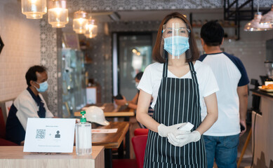Asian waitress woman wearing face masks and holding an infrared forehead thermometer to check body temperature for virus symptoms of customers before entering the restaurant ( coffee shop ).