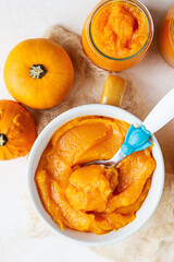 Homemade pumpkin puree in bowl with baby spoon and fresh pumpkins on light concrete background. The concept of baby food.