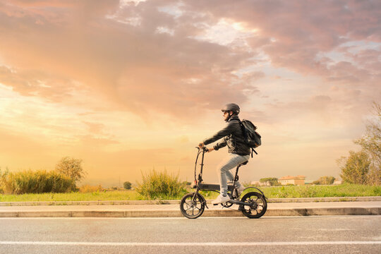 Smiling man with helmet and his electric bicycle at sunset. Commuter and new transportation concept. Green energy. Caucasian man drives his foldable e bike to go work or in leisure
