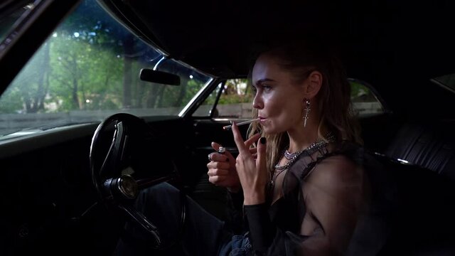 pretty lady is lighting cigarette and smoking sitting inside classic american car on driver seat