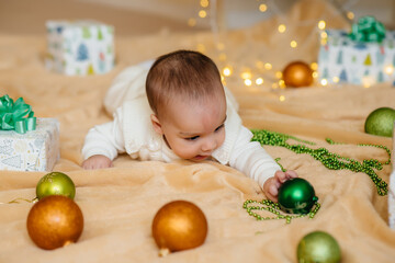 Fototapeta premium Cute smiling baby is lying under a festive Christmas tree and playing with gifts. Christmas and New Year celebrations