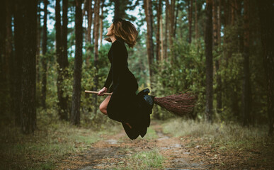 Fototapeta premium Young beautiful and mysterious Witch woman Flying on the Broom in woods. Levitation, a witch on a broom, free space. Halloween holiday, magic