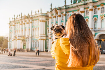 A young female owner in a yellow jacket holds a pug in her arms on a square in Saint Petersburg, tourist banner