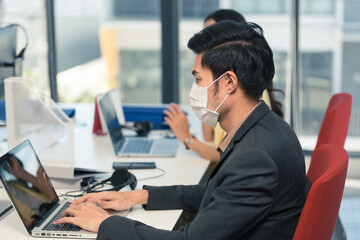 Asian male employee and woman co worker wearing face mask to protect virus infection typing on laptop computer at office desk. new normal working and social distancing concept. copy space