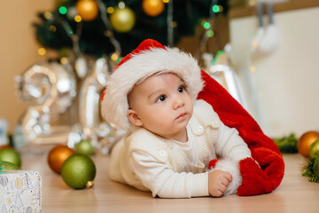 Cute smiling baby is lying under a festive Christmas tree and playing with gifts. Christmas and New Year celebrations