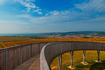 A round footbridge to the lookout tower in Kobyla in Moravia in the Czech Republic. There is a view of the countryside from the lookout tower. In the background is a blue sky with white clouds.