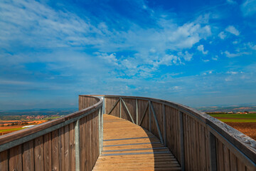 Fototapeta premium A round footbridge to the lookout tower in Kobyla in Moravia in the Czech Republic. There is a view of the countryside from the lookout tower. In the background is a blue sky with white clouds.