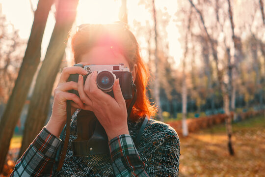 Person with vintage camera in autumn park. Portrait of a young beautiful woman freelancer in an autumn park with a photo camera. Red-haired girl journalist.