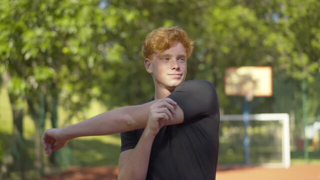 Confident young sportsman warming up before training outdoors and leaving. Middle shot portrait of smiling Caucasian redhead man stretching neck and hands on sunny outdoor court and walking away.