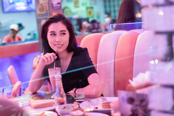 Young beautiful retro girl sitting in a diner restaurant. Woman in black eating in a diner with beautiful neon light. Pretty happy young woman sitting in the diner. Fashion girl in a vintage cafe.