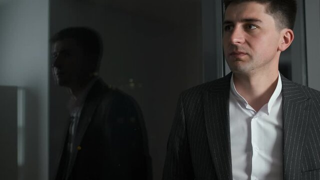 30-year-old businessman in a stylish suit stands in his office near a panoramic window overlooking the night city. He thinks about a business plan and drinks coffee and tea