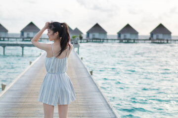 Beautiful traveler woman in a beach dress walks over a wooden pier on a tropical island. Young woman in white clothes walking on a wooden pier. Romantic honeymoon getaway in overwater bungalows villa.