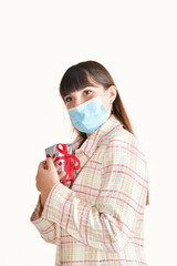 Close up of a young woman using a surgical mask holding a gift box close to her chest on a light background. Gifts and safety concept.