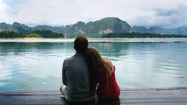 Back View of Happy Romantic Couple Sitting Together on Bridge and Woman Putting Head on Shoulder in Nature. Pair of Travel People on Vacation on Green Mountains Lake with Cloud Sky in Thailand