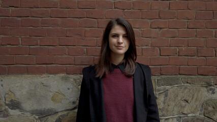 Portrait of cute young shy woman standing against red brick wall looking around and smiling for camera