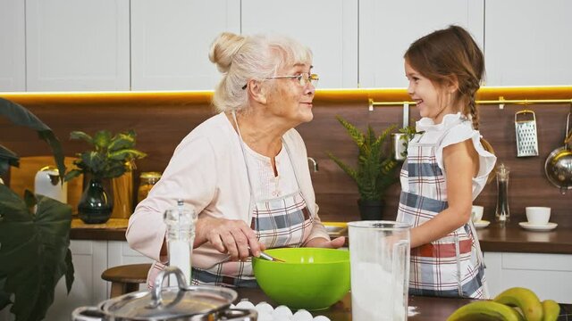 Grandmother is whipping eggs and her little granddaughter is staining grannys nose in flour, they going to bake cookies at modern kitchen