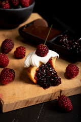 Muffin de mora cubierto de crema chantilly partido por la mitad sobre tabla y fondo negro al lado de cobertura de chocolate.