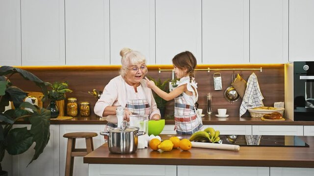 Grandmother is whipping eggs and her little grandchild is staining grannys nose in flour, they going to bake cookies at modern kitchen