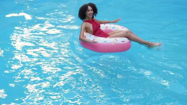 Alone young African female in red swimsuit swimming, pull, oar in pool in inflatable circle pink donut laughing looking at camera. Ethnicity Afro woman relax in blue water with rubber ring tube