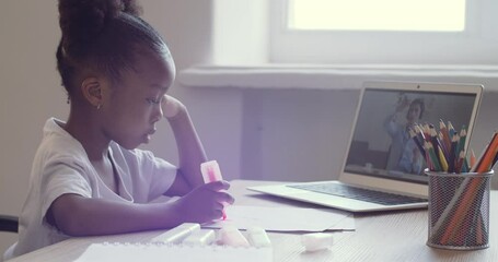 Cute little schoolgirl draws on paper sitting at desk looking into laptop screen. African American ethnic child watching video online speak with teacher remotely via webcam, answer tutor, e-learning - Powered by Adobe