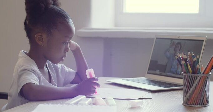 Cute little schoolgirl draws on paper sitting at desk looking into laptop screen. African American ethnic child watching video online speak with teacher remotely via webcam, answer tutor, e-learning