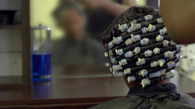 Blurred reflection in mirror of female African hair stylist combing client's hair to for perm rod at hair salon.