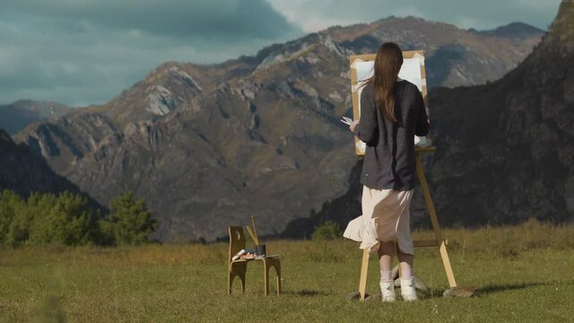 Beautiful girl paints a picture in the mountains. A professional artist creates a landscape on a molbert. Hobbies, hobbies and work in the fresh air.