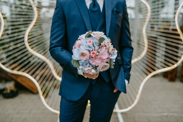 Groom in dark blue wedding suit with elegant round bride bouquet in his hands. Metall big ceremony arch on background and green glas house.