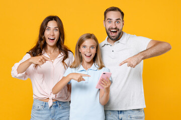Obraz premium Surprised funny young parents mom dad with child kid daughter teen girl in basic t-shirts pointing index fingers on mobile cell phone isolated on yellow background studio portrait. Family day concept.