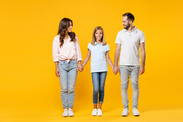 Fototapeta premium Full length portrait of smiling young parents mom dad with child kid daughter teen girl in t-shirts holding hands looking at each other isolated on yellow background. Family day parenthood concept.