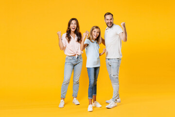 Obraz premium Full length portrait happy joyful laughing young parents mom dad with child kid daughter teen girl in t-shirts clenching fists doing winner gesture isolated on yellow background. Family day concept.