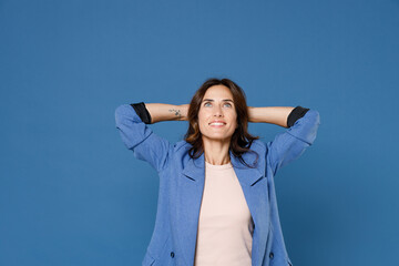 Smiling cheerful pensive beautiful attractive young brunette woman 20s wearing basic jacket looking up with hands behind head isolated on bright blue colour wall background, studio portrait.