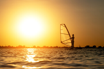 A beginner windsurfer woman stands on a board with a sail on a sunset background. Windsurfing school