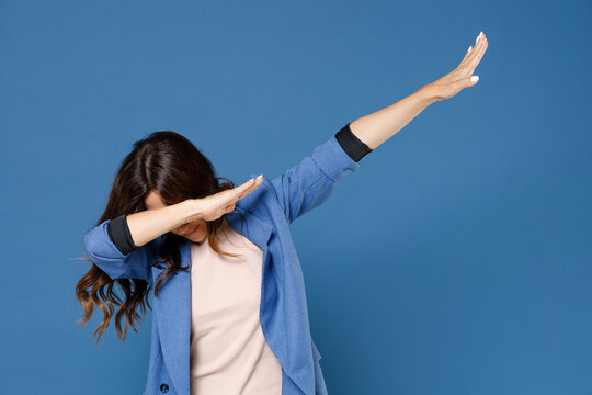 Cheerful funny young brunette woman 20s in basic jacket standing doing dab hip hop dance hands gesture, youth sign hiding and covering face isolated on bright blue colour background, studio portrait.