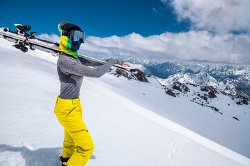 Close-up young woman skier in a ski mask with a closed face holds skis on her shoulder against the backdrop of snow-capped mountains