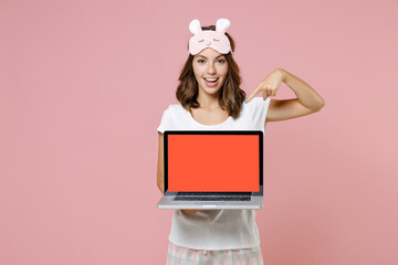 Funny young woman in white pajamas home wear sleep mask pointing index finger on laptop pc computer with blank empty screen resting at home isolated on pastel pink background. Relax good mood concept.