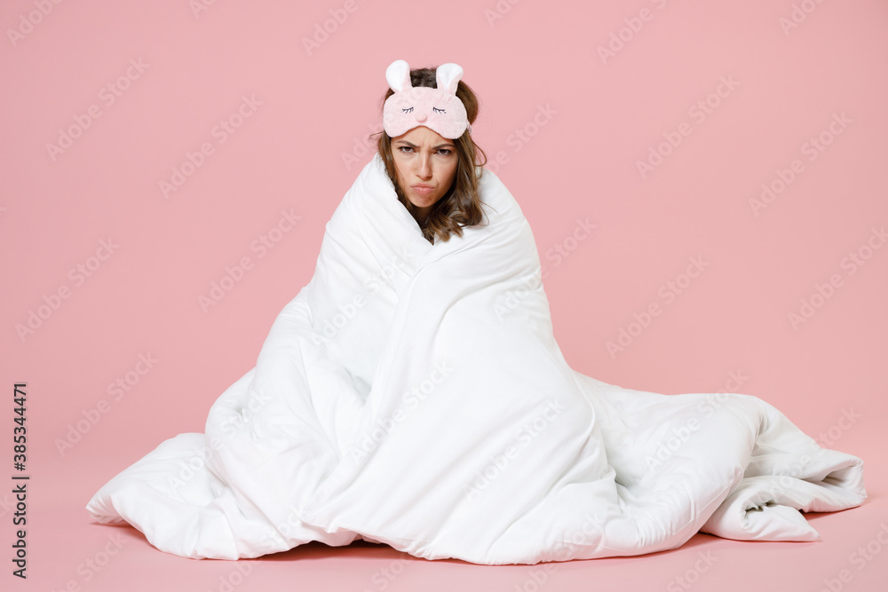 Wall mural Displeased tired angry young woman in pajamas home wear sleep mask sit wrapping in blanket looking camera rest at home isolated on pink background studio portrait. Relax good mood lifestyle concept.