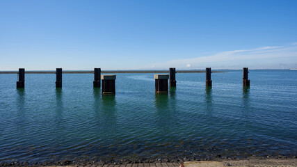 Dark wooden columns stand in the deep blue water, blue sky with white clouds, Dam in the foreground. Netherlands, Zeeland.