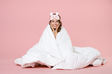 Smiling young woman in pajamas home wear sleep mask sitting wrapping in blanket put hands on cheeks...