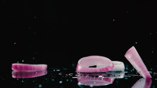 Ruedas de cebolla morada cayendo en camara lenta sobre una lamina negra con agua haciendo splash. HD
