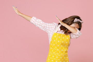 Young woman housewife in apron doing dab hip hop dance hands gesture youth sign hiding and covering face while doing housework isolated on pastel pink background studio portrait. Housekeeping concept.